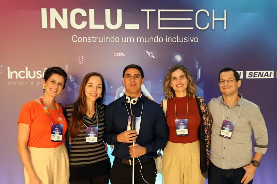 A imagem mostra um grupo de cinco pessoas posando para uma foto em frente a um fundo roxo com o texto "INCLU-TECH Construindo um mundo inclusivo" e o logotipo do SENAI. 