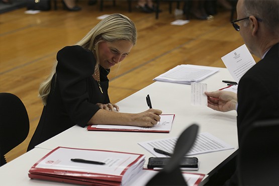Uma mulher loira vestida de preto está assinando um documento. A mesa em que está apoiada para assinar é branca, e muitos outros papéis e pastas vermelhas estão sobre ela. Um homem aparece no canto da imagem, e está alcançado um pequeno papel para a moça.