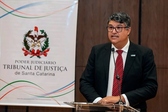 Um homem grisalho de terno preto e gravata vermelha com listras, fala no microfone em cima do palco. Atrás há um banner com a escrita "Poder Judiciário, Tribunal de Justiça de Santa Catarina".