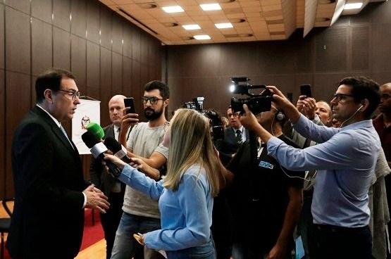 Muitos jornalistas entrevistam, gravam e tiram fotos de um participante do evento que está posicionado de pé, é alto, usa terno preto e óculos.