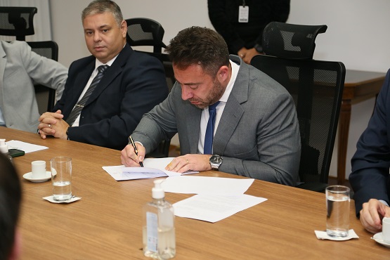 Dois homens estão numa sala, sentados em uma cadeira que está na lateral de uma mesa, um deles está com a mão cruzada, e outro está escrevendo sobre uma folha de papel.