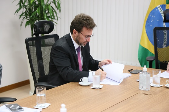 Foto da parte de uma mesa em que está um homem sentado em uma cadeira, escrevendo sobre um papel. Sobre a mesa, também estão alguns copos de água e alguns pertences pessoais. Ao fundo está uma bandeira do brasil e uma planta.