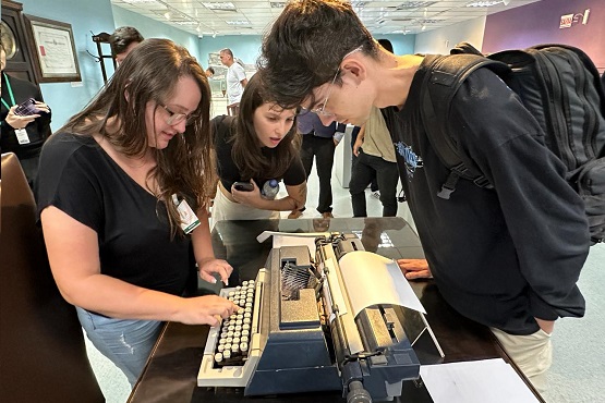 Três jovens ao redor de uma máquina de escrever, que está em cima de uma mesa de madeira. Menina à esquerda escreve na máquina de escrever, menina ao centro da imagem e menino à direita observam.