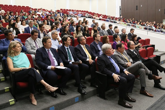 Fotografia de pessoas assistindo palestra em auditório com as cadeiras vermelhas acolchoadas. Os homens usam terno e as mulheres usam traje social.