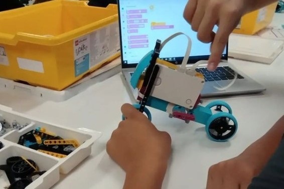 Em cima de uma mesa com tampo branco está um robô azul construído com peças e blocos de montar. Duas mãos mexem no brinquedo. Ao lado esquerdo da imagem está uma bandeja com peças de montar. No fundo à direita está um computador.
