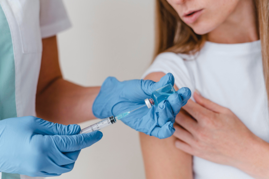 No primeiro plano, uma pessoa com luvas azuis pega o líquido dentro de uma ampola com um seringa de vacina. Ao fundo, uma outra pessoa arregaça a manga de uma blusa branca para tomar a vacina.