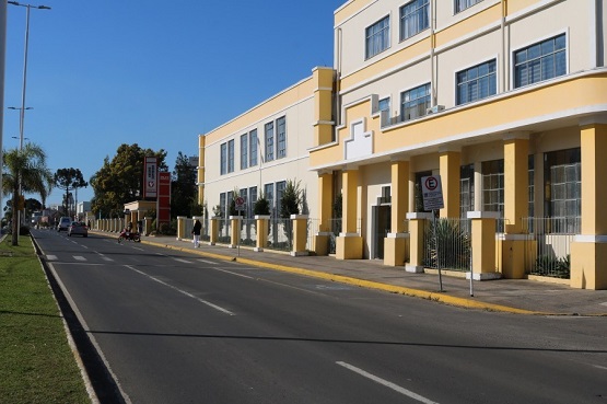A imagem mostra uma rua com um prédio amarelo de dois andares. O prédio tem várias janelas e uma entrada com colunas. À esquerda, há uma calçada com grama e algumas árvores.