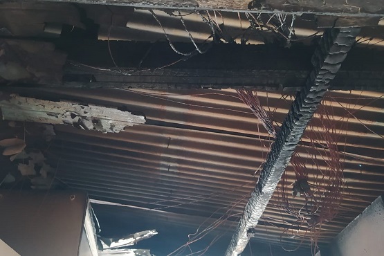 A imagem mostra o teto de uma estrutura que foi danificada por um incêndio. As vigas de madeira estão queimadas e há fios elétricos expostos e derretidos. O telhado de metal ondulado também está parcialmente destruído, com buracos e partes faltando.
