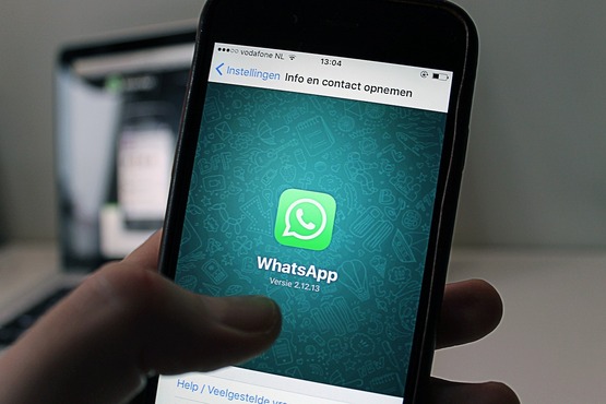 A imagem mostra uma mão segurando um celular. Na tela do celular, o aplicativo WhatsApp está aberto, exibindo o logo verde com o ícone de um telefone branco no centro. Ao fundo, há uma tela de computador desfocada.