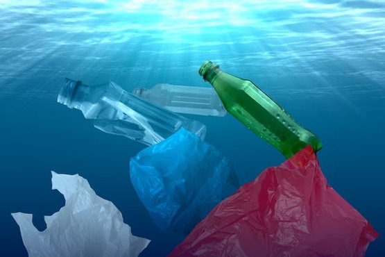 A imagem mostra o fundo no mar iluminado pela luz do sol. Dentro da água tem 3 garrafas plásticas, verde e transparente. Além de ter também 3 pedaços de sacolas plásticas, azuis vermelhas e brancas.
