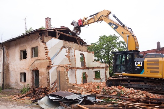 A imagem mostra uma escavadeira amarela da marca CAT demolindo um prédio antigo e deteriorado. O prédio parece estar em ruínas, com partes das paredes e do telhado já desmoronadas. Há entulho espalhado ao redor da área de demolição.