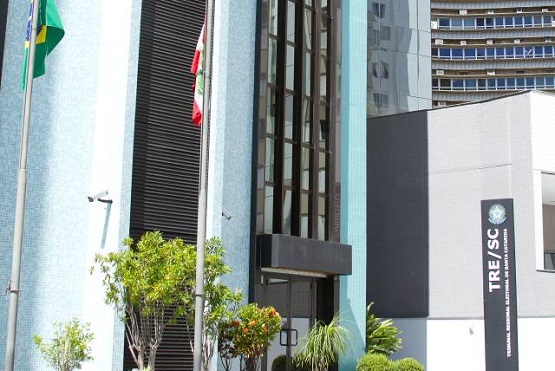 A imagem mostra a fachada de um prédio com a placa "TRE/SC" que significa Tribunal Regional Eleitoral de Santa Catarina. Há algumas árvores e plantas na frente do prédio, e a arquitetura parece moderna com linhas retas e vidros.