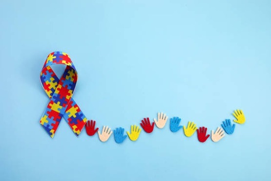 A imagem mostra um laço colorido com peças de quebra-cabeça, que é um símbolo frequentemente associado ao autismo. Abaixo do laço, há uma sequência de pequenas mãos de papel em várias cores (vermelho, azul, amarelo e bege), formando uma linha vertical. O fundo da imagem é azul claro.