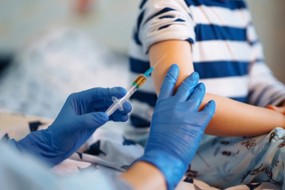 A imagem mostra uma pessoa recebendo uma injeção no braço. A pessoa que está aplicando a injeção está usando luvas azuis. O braço da pessoa que está recebendo a injeção está estendido, e ela está vestindo uma roupa com listras azuis e brancas.