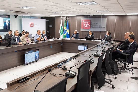 A imagem mostra uma reunião formal em uma sala de conferências. Há várias pessoas sentadas ao redor de uma mesa retangular grande. Na mesa, há microfones e cabos. Na parede ao fundo, há bandeiras do Brasil e de São Paulo, além de um painel com o logotipo do Ministério Público de São Paulo (MPSP).