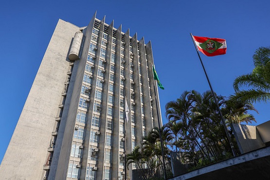 A imagem mostra o prédio do tribunal de justiça. Há uma bandeira de Santa Catarina hasteada e algumas árvores em volta. A construção tem muitas janelas e andares. O céu está azul.
