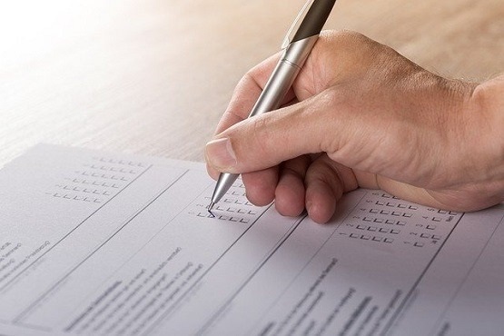 A imagem mostra uma mão segurando uma caneta preta assinalando quadrinhos em um papel que parece ser um documento. O papel está em cima de uma mesa de madeira, onde a mão da pessoa também está apoiada.