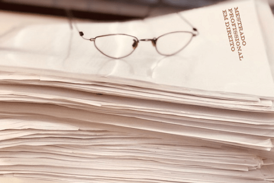 A imagem mostra uma pilha de papéis ou documentos à esquerda e um par de óculos de leitura descansando sobre um dos documentos à direita. No documento onde os óculos estão apoiados, está escrito "MESTRADO PROFISSIONAL EM DIREITO".