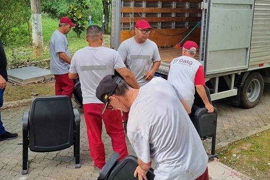 A imagem mostra cinco homens trabalhando ao lado de um caminhão de mudanças. Eles estão vestindo uniformes com calças vermelhas e camisetas cinza. Três deles também estão usando bonés vermelhos. Eles estão carregando cadeiras pretas, aparentemente colocando no caminhão. Ao fundo, há uma área verde com árvores e vegetação.